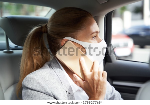 Business during coronavirus. Business woman wearing\
protective face mask talking by mobile phone while sitting on back\
seat in the car