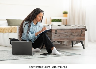 Business And Education Concept. Focused young woman sitting on floor leaning on bed working on laptop writing letter in paper notebook, free copy space. Millennial female studying at home, using pc