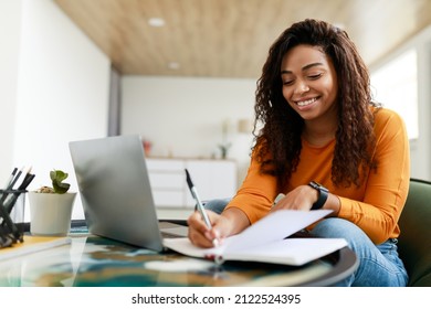 Business And Education Concept. Smiling young black woman sitting at desk working on laptop writing letter in paper notebook, free copy space. Happy millennial female studying using pc - Shutterstock ID 2122524395