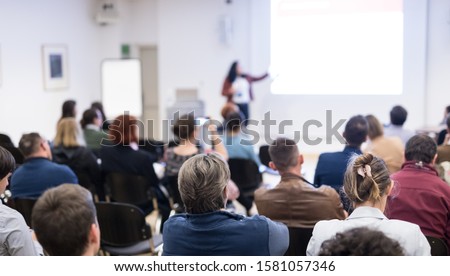 Business and entrepreneurship symposium. Female speaker giving a talk at business meeting. Audience in conference hall. Rear view of unrecognized participant in audience. Copy space on whitescreen.