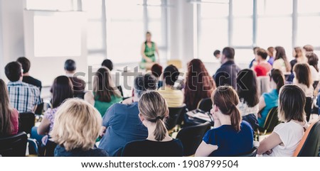 Business and entrepreneurship symposium. Female speaker giving a talk at business meeting. Audience in conference hall. Rear view of unrecognized participant in audience.