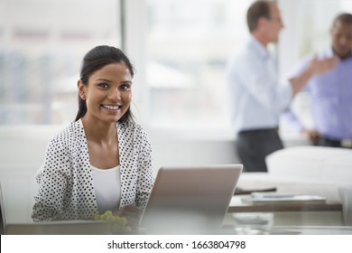 A business environment, a light and airy workplace in the city. A woman sitting at a desk using a laptop computer. Two men in the background. - Shutterstock ID 1663804798
