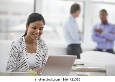 A Business Environment, A Light And Airy Workplace In The City. A Woman Sitting At A Desk Using A Laptop Computer. Two Men In The Background.