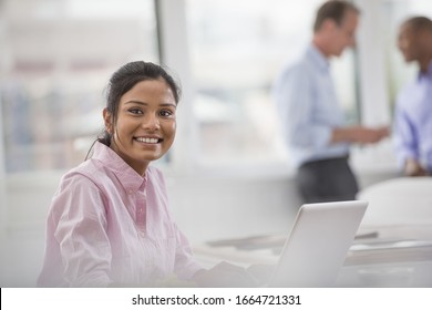 A Business Environment, A Light And Airy Workplace In The City. A Woman Sitting At A Desk Using A Laptop Computer. Two Men In The Background.