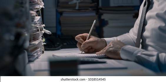 Business Executive Working Overtime At Night In The Office, He Is Writing Notes And Calculating Costs