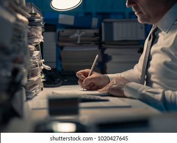 Business Executive Working Overtime At Night In The Office, He Is Writing Notes And Calculating Costs