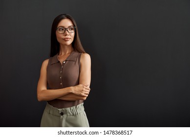 Business Expert. Young Asian Woman In Eyeglasses Keeping Arms Crossed And Looking Aside While Standing Against Dark Background