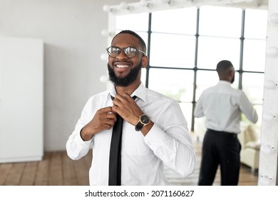 Business And Fashion Concept. Portrait Of Smiling Confident Young African American Businessman In Eyelasses Adjusting Necktie At Home, Getting Ready For Work, Wearing White Shirt And Luxury Watch