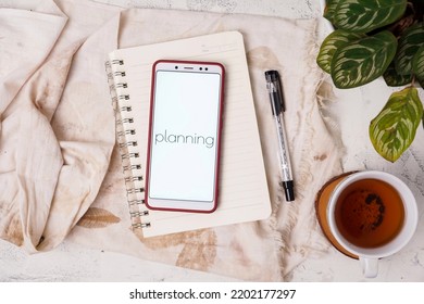 Business And Finance Concepts. The Screen Display On The Smartphone Reads Planning. Notebook Background, Vintage Look With A Glass Of Warm Tea. Top View.