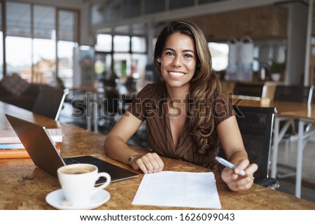 Business, finance and entrepreneur concept. Happy cheerful attractive spanish woman sitting alone cafe, co-working space with opened laptop, working remote, drink coffee and read paper, studying