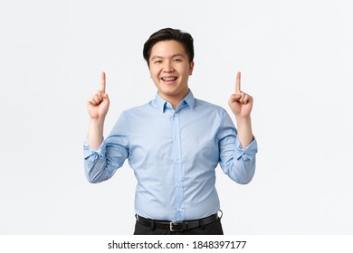 Business, Finance And People Concept. Upbeat Good-looking Asian Businessman In Blue Shirt, Pointing Fingers Up And Smiling Happy, Showing Announcement, Salesman Recommend Product