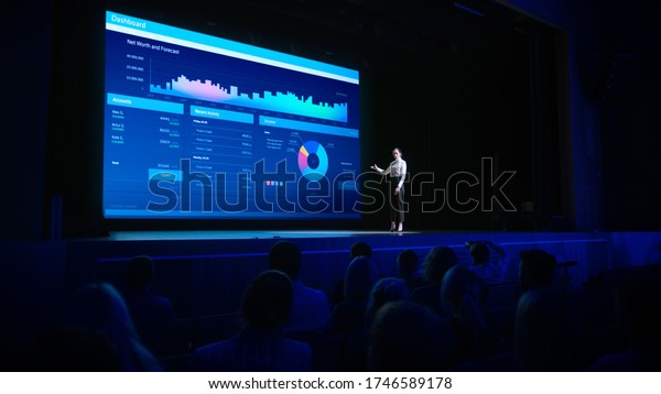 Business Forum Economics Conference Stage: Visionary\
Female Chief Analyst Delivers Speech and Shows Infographics,\
Statistics on Movie Theater Screen. Presentation with Speaker in\
Full Auditorium Hall