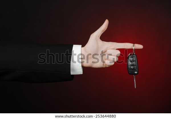 Business and\
gift theme: car salesman in a black suit holds the keys to a new\
car on a dark red background in\
studio