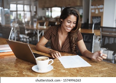 Business, Gig Economy Concept, Charming Young Brazilian Female Entrepreneur Sitting Co-working Cafe, Reading Working Paper, Taking Notes, Using Laptop As Working Remote From Office, Drink Cappuccino