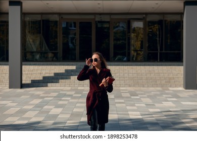 A Business Girl In A Burgundy Coat And Black Glasses Walks Away From The Business Center With A Notebook On A Clear, Sunny Day. A Student With A Textbook Is Walking Near An Educational Institution Wea