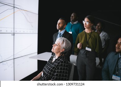 Business Group Having A Discussion During A Conference, Looking A Large Video Wall With Charts And Graphs