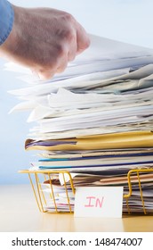 Business image of a male hand with blue shirt cuff visible, adding or removing document from tall pile in overflowing office In tray.
