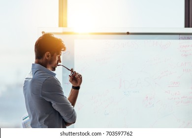 Business investor in deep thought looking at the business ideas written on the whiteboard. Businessman thinking while holding his spectacles to mouth - Shutterstock ID 703597762
