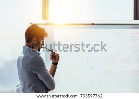 Business investor in deep thought looking at the business ideas written on the whiteboard. Businessman thinking while holding his spectacles to mouth