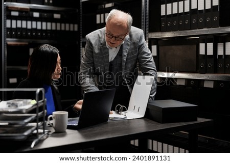 Business invoicing management employees comparing accountancy market data analysis between laptop and paperwork. Archival depository filled with document folders on cabinet shelves and graphic charts