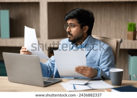 Business Issue. Concerned Indian Businessman Holding Papers Reading Bad News, Having Problems With Financial Bills Sitting At Desk With Laptop In Modern Office Indoor. Entrepreneurship Difficulties