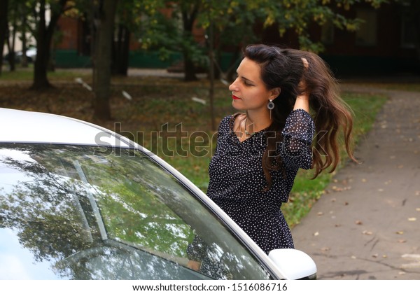 Business lady in an elegant dress\
straightens her hair standing next to a white car on the street\
background.Concept: successful\
woman.Business