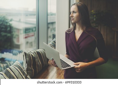 Business Lady In Purple Suit Is Standing In Office Settings Next To Window Of Chill Out Area, Holding Laptop And Looking Outside With Copy Space Zone For Advert, Message Or Your Logo