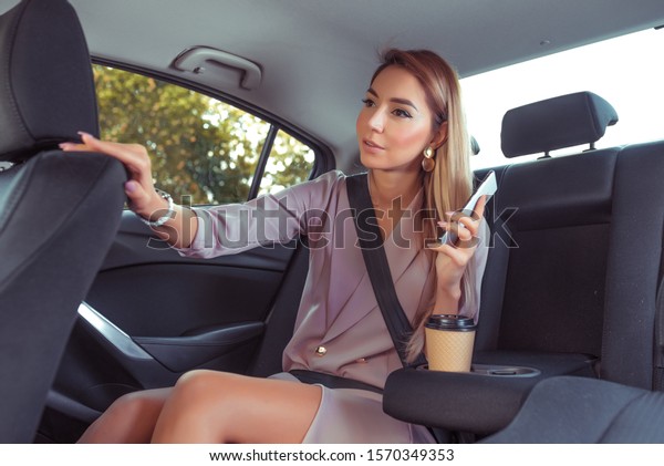 Business lady woman in taxi car cabin, summer day\
city, tells taxi driver route, asks to stop, talks on smartphone,\
specifies arrival route. Casual makeup pink formal suit. A cup of\
coffee with tea
