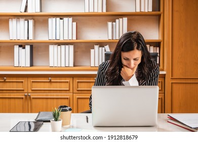 A business or lawyer woman in a black stripe suit putting a hands on chin working in front of a laptop computer in an office room with a bookshelf in the background. Serious thinking focus expression.