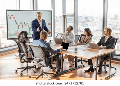 A business leader presents financial data to his team during a meeting in a modern office. Leader stands near a large screen displaying a chart, while the team members sit at a table in front of him - Powered by Shutterstock