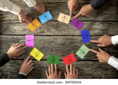 Business Leaders Creating Diverse And Competent Business Team Seated Around A Wooden Table Each Pointing To Two Brightly Colored Cards With Hand Doodled Person, Overhead Close Up Of Their Hands.