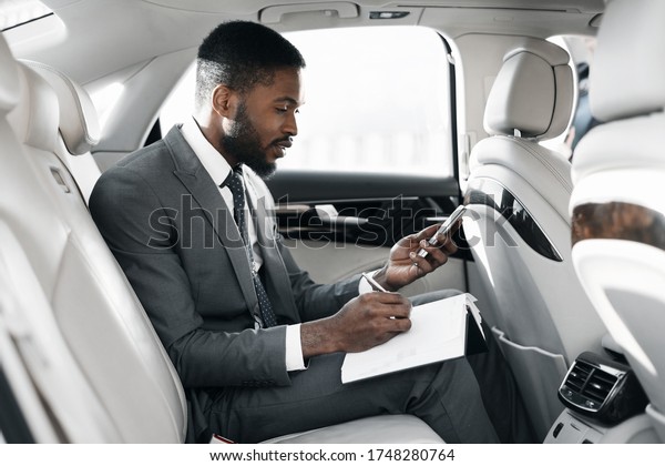 Business Lifestyle. Black Businessman Using\
Smartphone Taking Notes Making Work Schedule Sitting In Car On Back\
Seat. Selective Focus