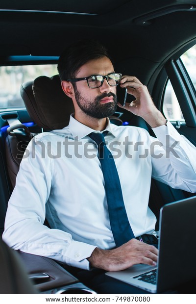 Business lifestyle.\
Handsome young man talking on mobile phone and using laptop\
while\
sitting in\
car\
