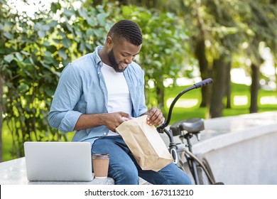 Business Lunch In Park. Cheerful African American Guy Unwrapping Package With Food, Resting Outdoors With Laptop During Break At Work