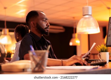 Business Man Analyzing Paperwork Report And Taking Notes To Create Research Presentation In Office With Big Windows. Working On Project Data With Documents And Computer During Sunset.