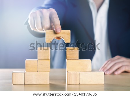 Business man bridging the gap between two towers or parties made from wooden blocks; conflict management or mediator concept, blue toned with ligth flare