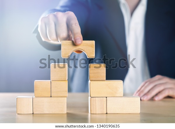 Business man bridging the gap between two\
towers or parties made from wooden blocks; conflict management or\
mediator concept, blue toned with ligth\
flare