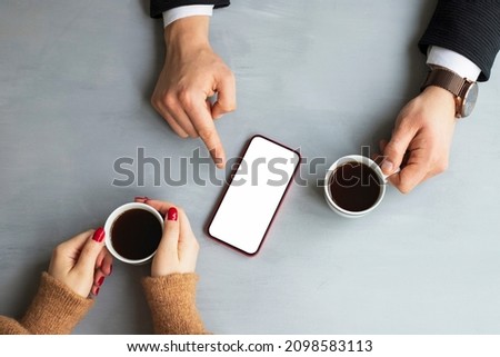 Business man in cafe showing blank phone screen to business woman with coffee cup on gray wooden table background. Top view.