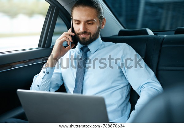 Business Man In Car Calling On
Phone While Going To Work. Handsome Smiling Young Male Talking On
Smartphone, Sitting On Back Seat Of Business Car. High Quality
Image.