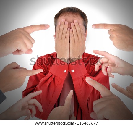 A business man is covering his eyes with his hands and has blame and shame with fingers pointing at him.
