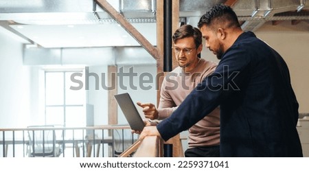 Business man discussing a tech project with his colleague in an office. Two business men using a laptop on an interior balcony. Male professionals working together.