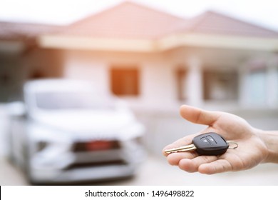 Business Man Hand Holding Car Keys Front With New Car On Background. Parking In Front Of The House. Transportation Concept. Leave Copy Space To Write Messages Text.