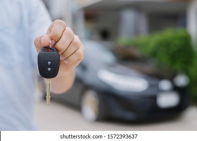 Business Man Hand Holding Car Keys Front With New Car On Background. Parking In Front Of The House. Transportation Concept. Leave Copy Space To Write Messages Text.