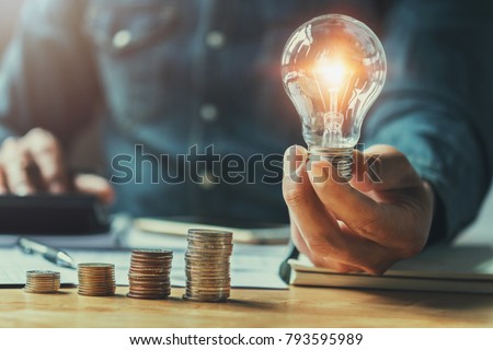business man hand holding lightbulb with using calculator to calculate and money stack. idea saving energy and accounting finance in office concept