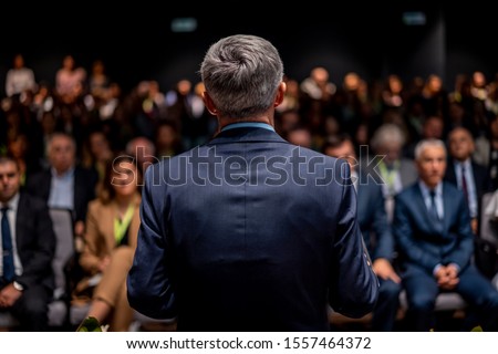 Business man is making a speech in front of a big audience at a conference hall. Speaker giving a talk on corporate business or political conference. Politician talking to group of people