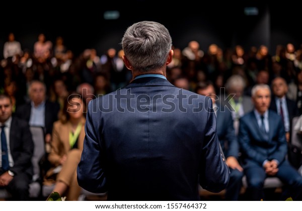 Business man is making a speech in front of a big
audience at a conference hall. Speaker giving a talk on corporate
business or political conference. Politician talking to group of
people