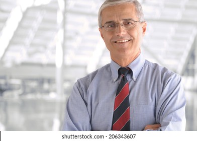 Business Man Portrait In Modern Office Building. Middle Aged Man Is Smiling At The Camera And Has His Arms Folded. Closeup Head And Shoulders Only.