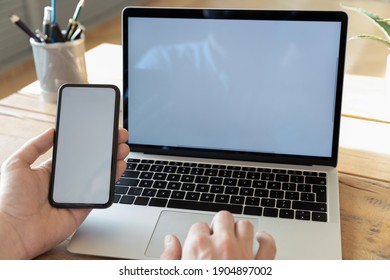 Business man sitting at desk using wireless computer holding smart phone with white empty mock up screen, close up view, multitasking, modern technology, new app installation, information sync concept