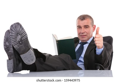 Business Man Sitting With His Legs On The Desk And Holding A Book While Showing The Thumb Up Gesture To The Camera. On A White Background