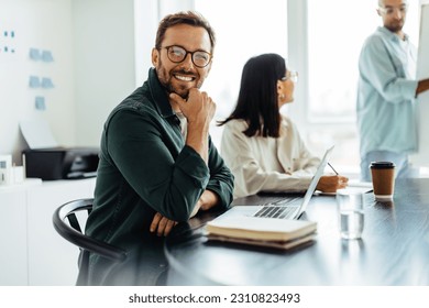 Business man smiling at the camera while sitting in a boardroom during a meeting. Happy business professional sitting in a presentation with a laptop. - Shutterstock ID 2310823493